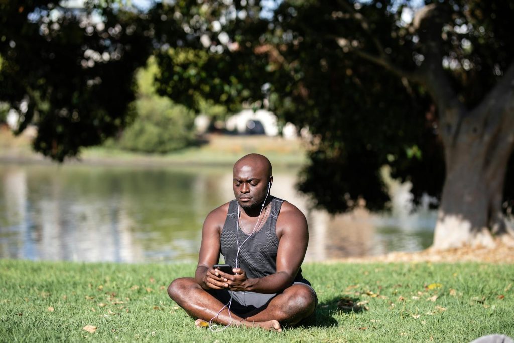 Adult man sitting cross-legged on grass, meditating with smartphone and earphones outdoors.