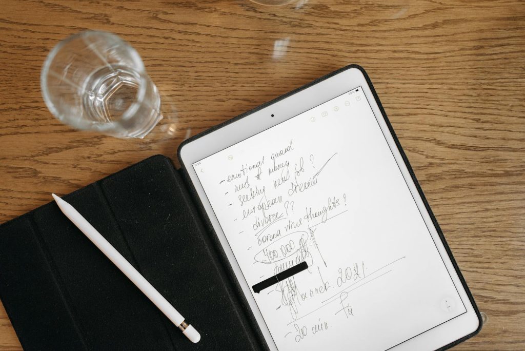 Top-down view of a tablet with handwritten notes, stylus, and glass on a wooden surface.