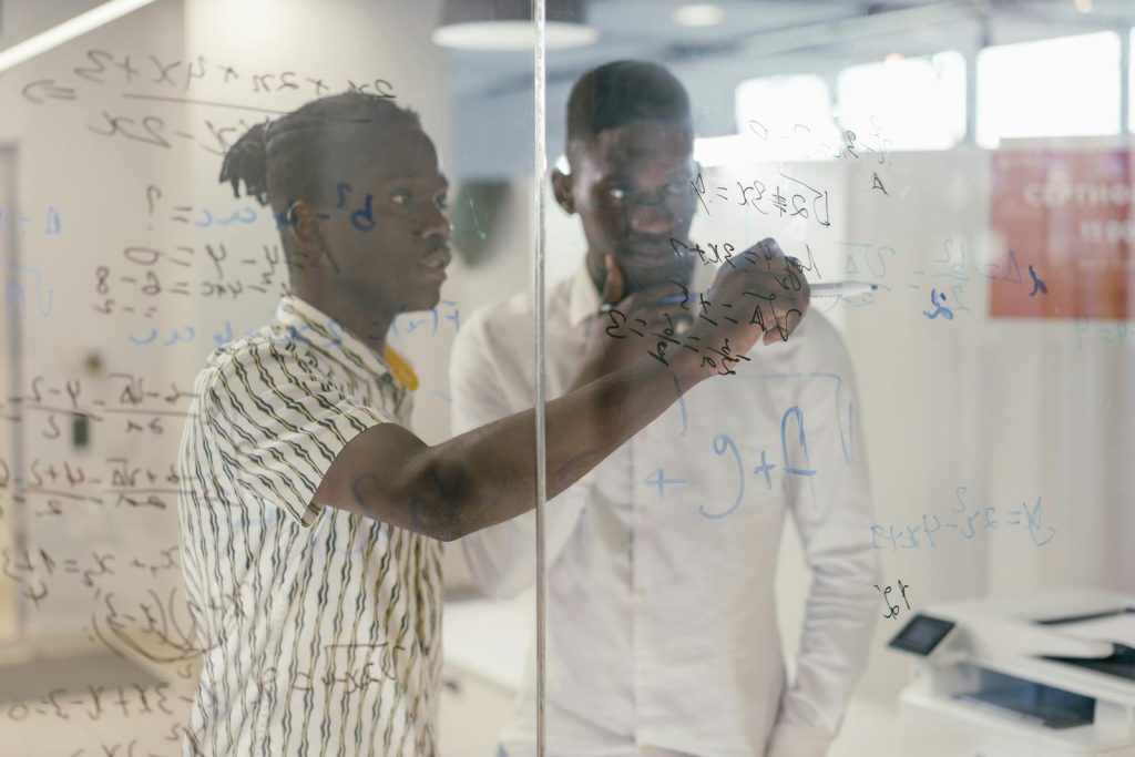 Two professionals discussing ideas and equations on a glass board in an office setting.