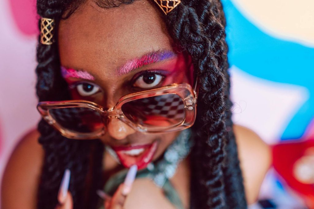 Vibrant portrait of a woman in sunglasses, featuring bold makeup and colorful eyebrows.